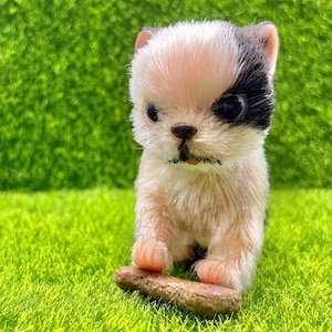 Peut inclure: Un petit chien en peluche blanc et noir avec un patch noir sur un œil. Le chien est assis sur une surface verte et tient un bâton brun dans ses pattes.