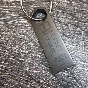 May include: A rectangular dark gray metal keychain with a circular split ring at the top. The surface features the engraved text AI CAN'T CREATE ART, with the letters arranged vertically and horizontally in a clean, sans-serif font style.