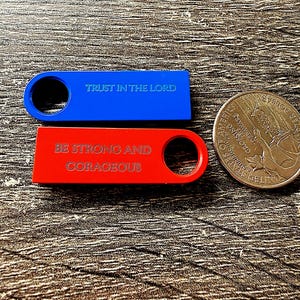 Könnte beinhalten: Ein blaues rechteckiges Schild mit einem kreisförmigen Loch, graviert mit dem Text TRUST IN THE LORD, und ein rotes rechteckiges Schild mit einem kreisförmigen Loch, graviert mit dem Text BE STRONG AND CORAGEOUS, neben einer US-Münze.