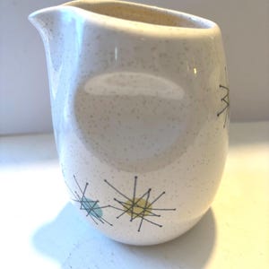 May include: A cream-colored ceramic creamer with a speckled finish and a spout. The exterior features a pattern of thin black lines forming starburst shapes, accented with small light blue and yellow circles scattered across the surface.