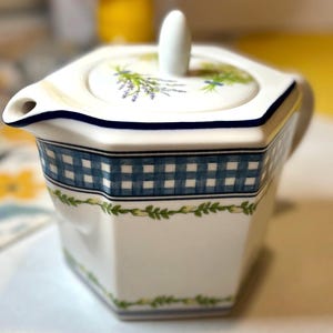 May include: White ceramic creamer with a blue and white checkered band and a lid featuring a lavender and floral design. The creamer has a spout and a white handle. The creamer is decorated with green leaf patterns.
