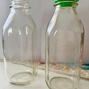 May include: Two clear glass milk bottles, one with a green plastic cap. The bottles have a classic square shape with rounded shoulders and a wide mouth. The bottles are empty and sitting on a white surface.
