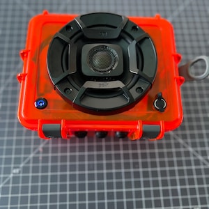 May include: A bright orange waterproof speaker with a black speaker grill and the word "Polk" printed on it. The speaker has a blue power button and a black button on the side.