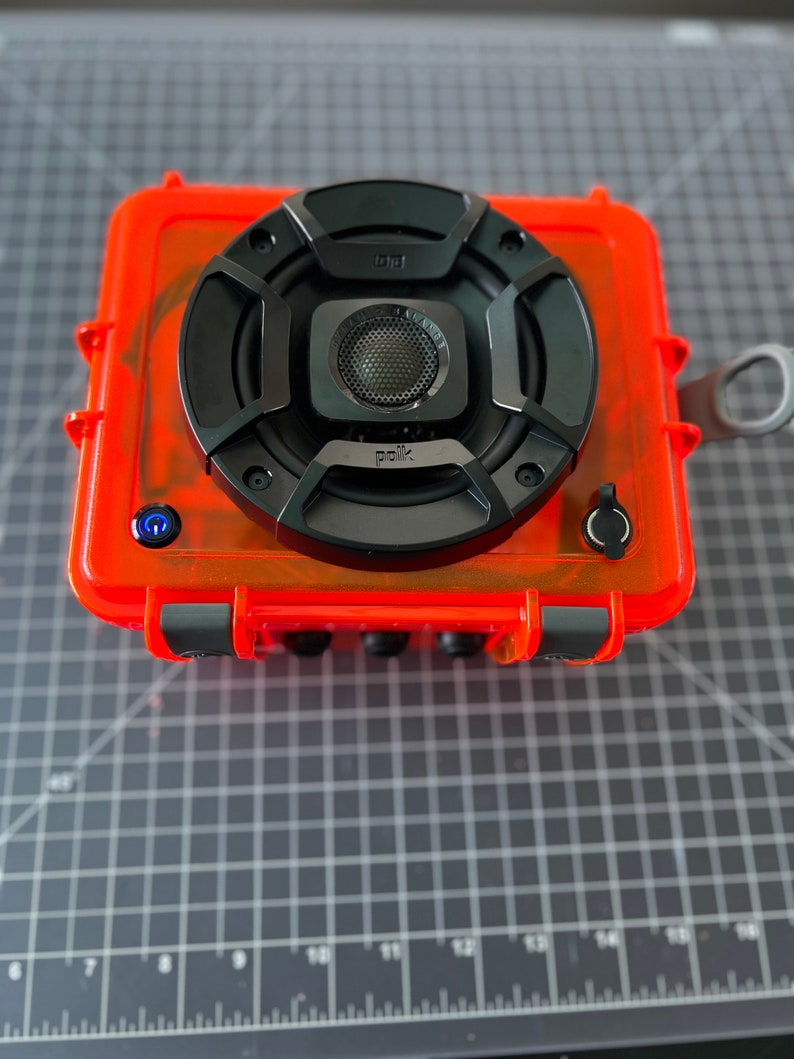 May include: A bright orange waterproof speaker with a black speaker grill and the word "Polk" printed on it. The speaker has a blue power button and a black button on the side.
