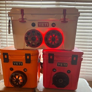 May include: Three Yeti coolers stacked on top of each other. The top cooler is white with red accents and speakers with red lights. The middle cooler is orange, and the bottom cooler is red. Each cooler has the "Yeti" logo.