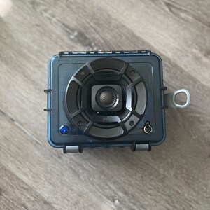 May include: A black and blue waterproof speaker with a Polk Audio logo and a blue light indicator. The speaker has a round, black speaker grille with a silver center.