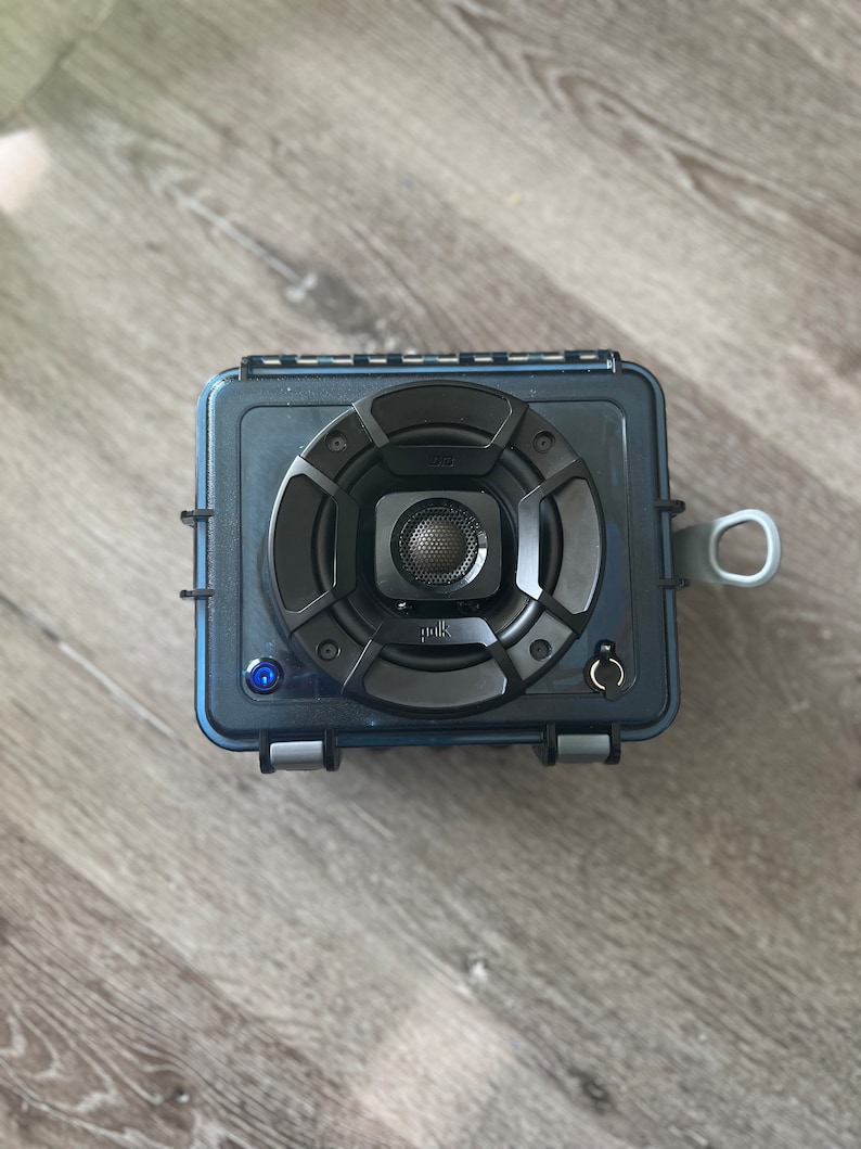 May include: A black and blue waterproof speaker with a Polk Audio logo and a blue light indicator. The speaker has a round, black speaker grille with a silver center.