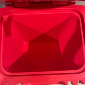May include: A bright red cooler with an open lid, revealing the empty interior. The cooler has a rectangular shape with rounded corners and a smooth, glossy finish. The interior is also red, with a slightly textured surface.