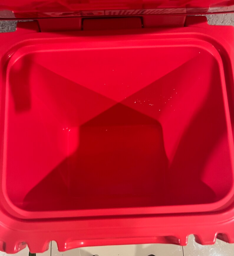 May include: A bright red cooler with an open lid, revealing the empty interior. The cooler has a rectangular shape with rounded corners and a smooth, glossy finish. The interior is also red, with a slightly textured surface.