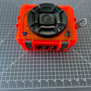 May include: A bright orange waterproof speaker with a black speaker grille and a black Polk Audio logo. The speaker is sitting on a gray cutting mat.