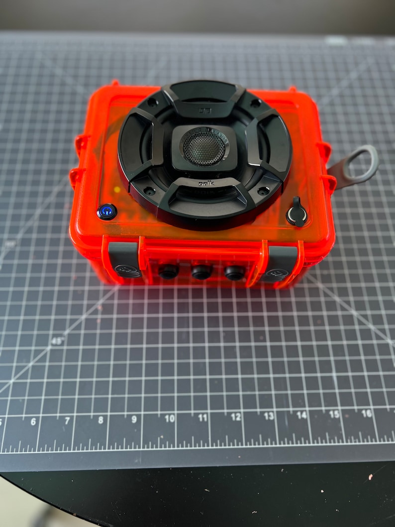 May include: A bright orange waterproof speaker with a black speaker grille and a black Polk Audio logo. The speaker is sitting on a gray cutting mat.