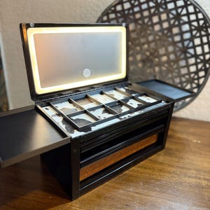 Könnte beinhalten: Schwarzer Schmuck-Organizer mit eingebautem beleuchteten Spiegel. Der Organizer hat mehrere Fächer und Schubladen mit einem Kuhfellmuster im Inneren. Der obere Deckel ist geöffnet und zeigt den Spiegel und die Ablageflächen.