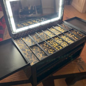 May include: Black makeup organiser with a built-in illuminated mirror. The interior features a leopard print design and multiple compartments for storage. The mirror is surrounded by bright white LED lights. The organiser is open, revealing the interior.