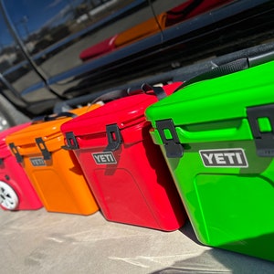 May include: A row of colorful YETI coolers in red, orange, and green. The coolers are rectangular with black latches and the YETI logo. One cooler has wheels. The coolers are lined up on a concrete surface.