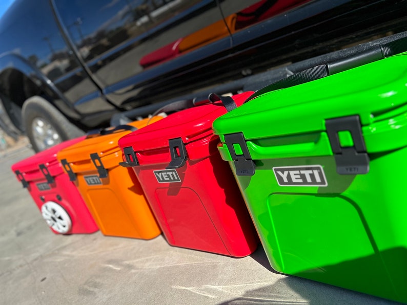 May include: A row of colorful YETI coolers in red, orange, and green. The coolers are rectangular with black latches and the YETI logo. One cooler has wheels. The coolers are lined up on a concrete surface.