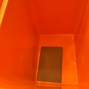 May include: An orange plastic bin with a rectangular base and a brown paper envelope inside. The envelope is folded and has text printed on it. The bin is empty and has a smooth, glossy finish.
