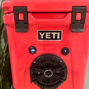 May include: A red YETI cooler with black accents, featuring a built-in speaker and voltage display. The cooler has a black speaker grill and a power button. The YETI logo is prominently displayed on the front.
