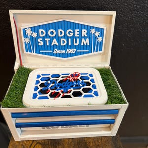 Könnte beinhalten: Eine weiße Box mit einem blau-weißen Dodger Stadium-Schild und einem blau-weißen Baseball-Motiv. Auf dem Schild steht "DODGER STADIUM Since 1962". Die Box hat eine grasartige Basis und blaue Akzente.