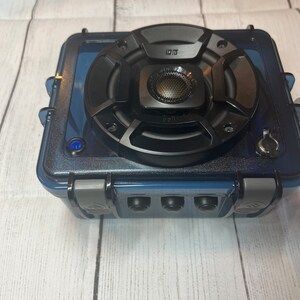May include: A black waterproof speaker with a blue case and a single button on the front. The speaker has a large black speaker driver and a smaller black speaker driver in the center. The speaker is sitting on a white wooden surface.