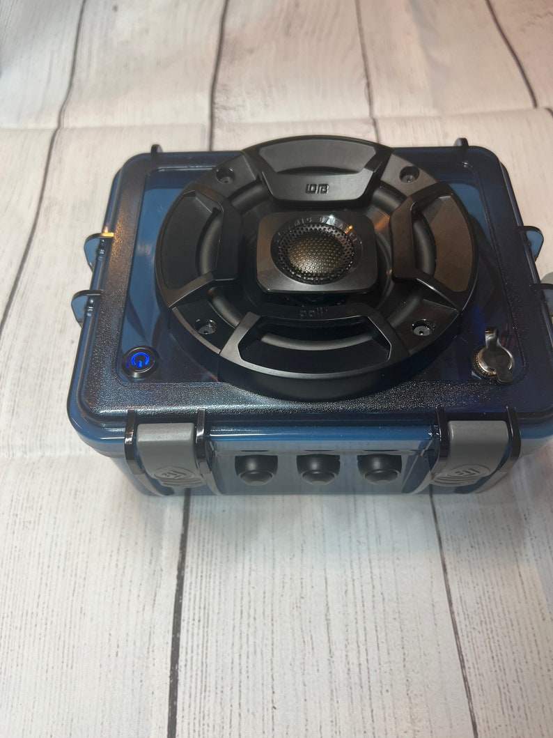 May include: A black waterproof speaker with a blue case and a single button on the front. The speaker has a large black speaker driver and a smaller black speaker driver in the center. The speaker is sitting on a white wooden surface.