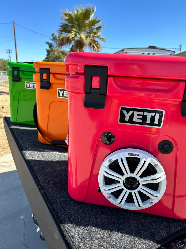 May include: Three colorful coolers: green, orange, and red. The red cooler features a Kicker speaker and a voltage control panel. Each cooler has a black handle and the Yeti logo.