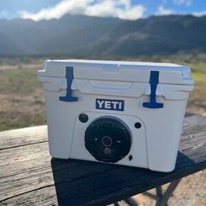 May include: White cooler with blue accents, featuring a built-in speaker. The cooler has the "YETI" logo and is set on a wooden table outdoors. The background includes mountains and a clear sky.