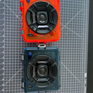 May include: Two waterproof portable speakers, one in a red case and one in a blue case. Both speakers have a black speaker grill with a smaller speaker in the center. The speakers are on a gray cutting mat.