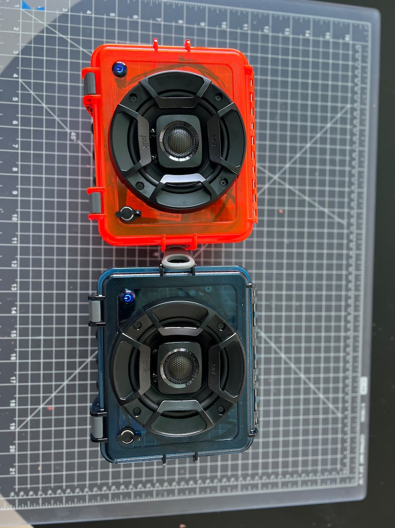May include: Two waterproof portable speakers, one in a red case and one in a blue case. Both speakers have a black speaker grill with a smaller speaker in the center. The speakers are on a gray cutting mat.