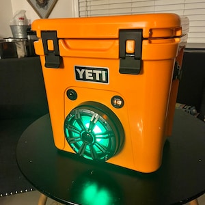 May include: An orange cooler with a black handle and a speaker with green lights. The cooler has the brand name "YETI" on the front. The speaker has the brand name "KICKER" on it.