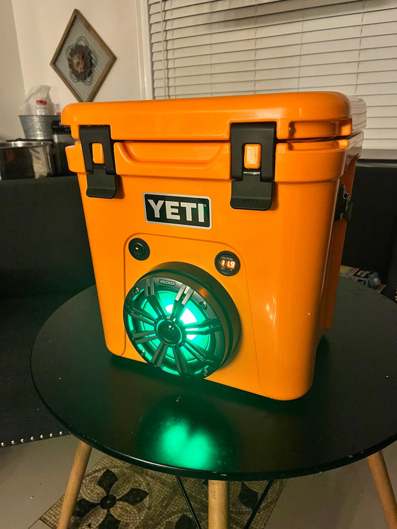 May include: An orange cooler with a black handle and a speaker with green lights. The cooler has the brand name "YETI" on the front. The speaker has the brand name "KICKER" on it.