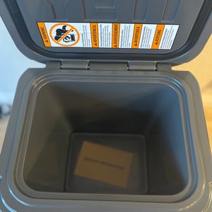 May include: A gray, open YETI cooler with a rectangular shape. The lid is open, revealing the interior. A warning label is visible on the inside of the lid. The cooler is sitting on a light brown surface.