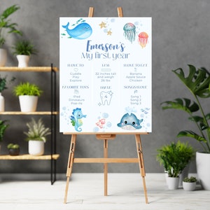 Könnte beinhalten: Ein blaues und weißes druckbares Poster mit einem Seepferdchen, einem Wal und einer Qualle. Das Poster ist für den ersten Geburtstag eines Kindes und enthält Informationen über das Alter, die Größe, das Gewicht, die Lieblingsspielzeuge und die Lieder des Kindes. Das Poster sagt "Emerson's My First Year".