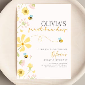 May include: A cream-colored invitation with floral and bee illustrations. The text reads "OLIVIA'S first bee day" and "Olivia's FIRST BIRTHDAY!" with event details. Yellow and pink flowers and bees decorate the corners.