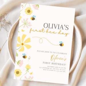 May include: A cream-colored invitation with floral and bee illustrations. The text reads "Olivia's first bee day" in a mix of gray and gold script. Additional text includes birthday details and RSVP information.