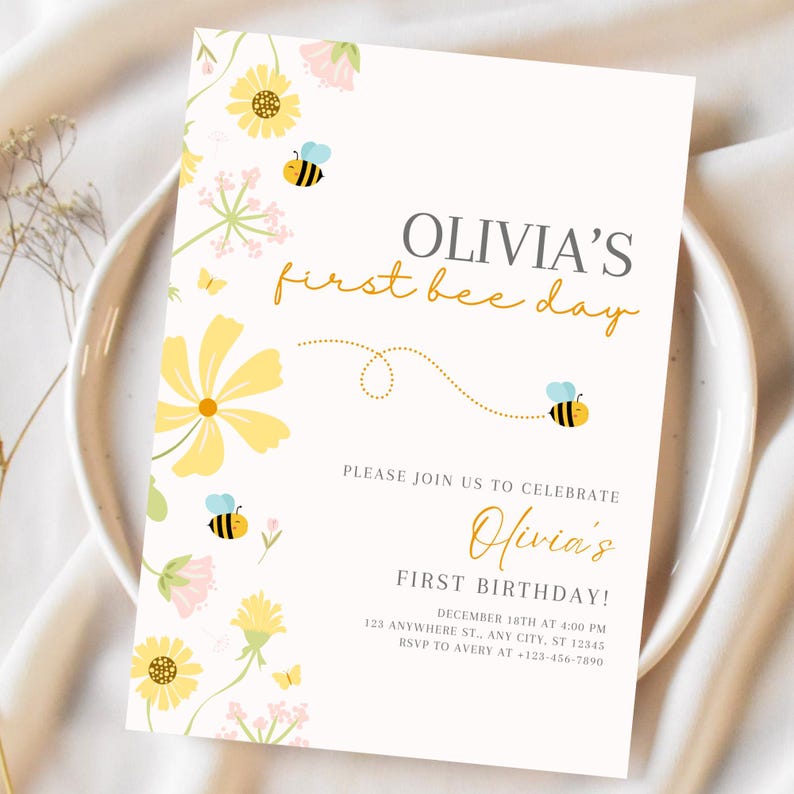 May include: A cream-colored invitation with floral and bee illustrations. The text reads "Olivia's first bee day" in a mix of gray and gold script. Additional text includes birthday details and RSVP information.