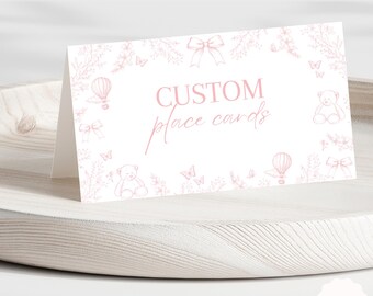 Op de afbeelding: Een witte, op maat gemaakte plaatskaart met roze bloemen- en teddybeerillustraties. De kaart bevat de tekst "CUSTOM place cards" in een sierlijk roze lettertype. De kaart wordt gepresenteerd op een licht houten dienblad.