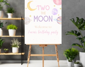 May include: A white sign with pink and gold lettering that says "Two the Moon" and "Welcome to Lucia's birthday party". The sign is decorated with stars, planets, and a crescent moon. The sign is on a wooden easel.