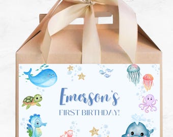 May include: A blue and white favor box with a sea-themed design. The box has a handle and is decorated with a whale, turtle, starfish, jellyfish, octopus, and seahorse. The text on the box reads "Emerson's First Birthday!"