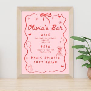 May include: A framed pink sign with the text "Olivia's Bar" in red script. The sign lists wine, beer, basic spirits, and soft drink options. Decorative elements include hearts, stars, and a bow.