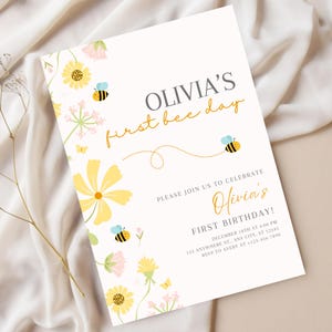 May include: A white invitation card with the text "OLIVIA'S first bee day" in gold and gray. The card is decorated with yellow flowers, pink accents, and cartoon bees. The invitation is for Olivia's first birthday.
