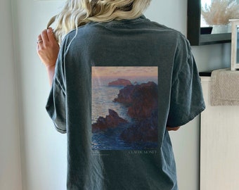 Può includere: Una maglietta grigio scuro con una stampa vintage di un dipinto di paesaggio marittimo di Claude Monet. Il dipinto raffigura una costa rocciosa con un cielo al tramonto e il testo "1886 Claude Monet" è stampato sotto l'immagine.