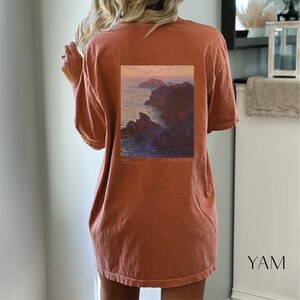 以下が含まれることがあります： クロード・モネの絵画がプリントされた焦げ茶色のTシャツ。絵画は、岩の多い崖と夕焼けの空が描かれた海の風景です。絵画の下には「1886」と「クロード・モネ」の文字がプリントされています。