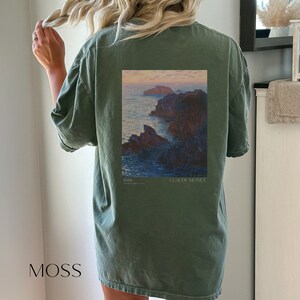 以下が含まれることがあります： クロード・モネの絵画がプリントされた緑色のTシャツ。絵画は、岩の多い崖と夕焼けの空が描かれた海の風景です。絵画の下には「1880」と「クロード・モネ」の文字がプリントされています。