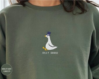 Könnte beinhalten: Ein grüner Pullover mit einer weißen gestickten Gans, die einen blauen Zylinder trägt. Der Text "Silly Goose" ist unter der Gans gestickt.