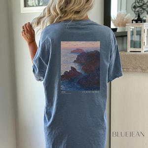 以下が含まれることがあります： クロード・モネの絵画がプリントされた青いTシャツ。絵画は、夕焼けを背景に岩だらけの海岸線を表しています。絵画の下には「1886」と「CLAUDE MONET」の文字が印刷されています。