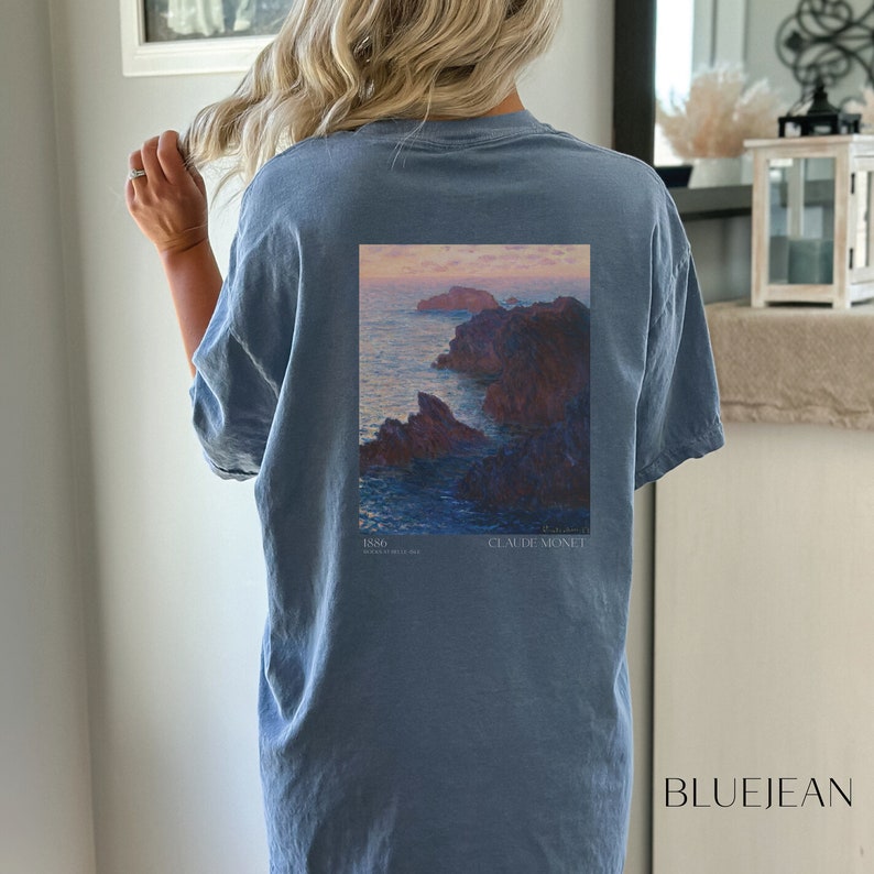 以下が含まれることがあります： クロード・モネの絵画がプリントされた青いTシャツ。絵画は、夕焼けを背景に岩だらけの海岸線を表しています。絵画の下には「1886」と「CLAUDE MONET」の文字が印刷されています。