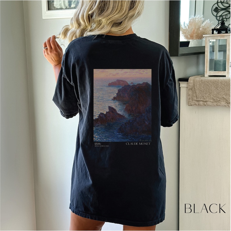 以下が含まれることがあります： 1886 年のクロード・モネの絵画「Coin de la plage, Belle-Isle」がプリントされた黒いTシャツ。絵画は、夕暮れ時の背景に岩だらけの海岸の風景を描いています。