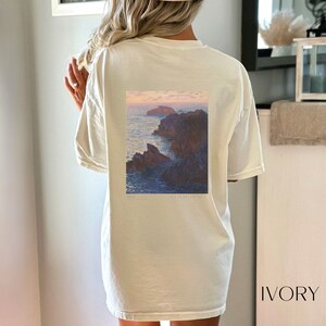 以下が含まれることがあります： クロード・モネの絵画がプリントされた、白のオーバーサイズTシャツ。絵画は、岩の多い崖と夕焼けの空が描かれた海の風景です。画像の下には「1886」と「CLAUDE MONET」の文字が印刷されています。シャツには「IVORY」のラベルが付いています。