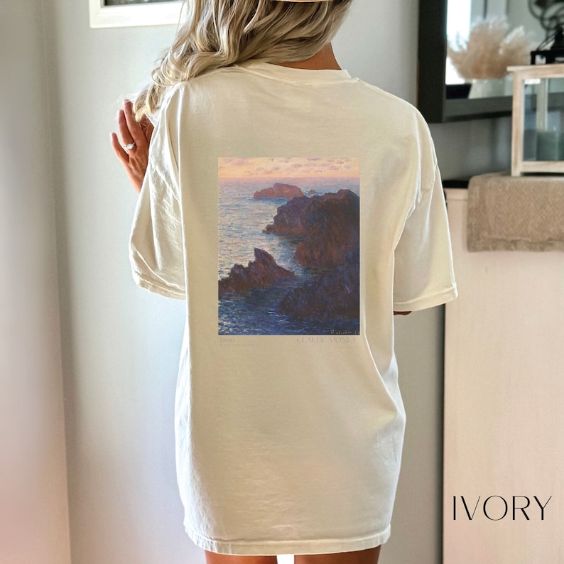 以下が含まれることがあります： クロード・モネの絵画がプリントされた、白のオーバーサイズTシャツ。絵画は、岩の多い崖と夕焼けの空が描かれた海の風景です。画像の下には「1886」と「CLAUDE MONET」の文字が印刷されています。シャツには「IVORY」のラベルが付いています。