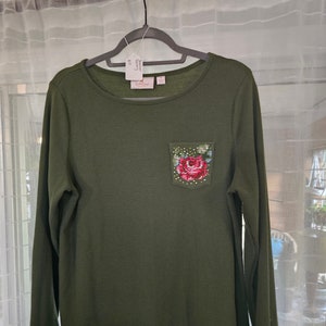 May include: Olive green long-sleeve shirt with a waffle knit texture. The shirt features a round neckline and a small pocket on the chest. The pocket is decorated with a cross-stitch floral design in shades of red, pink, and green.